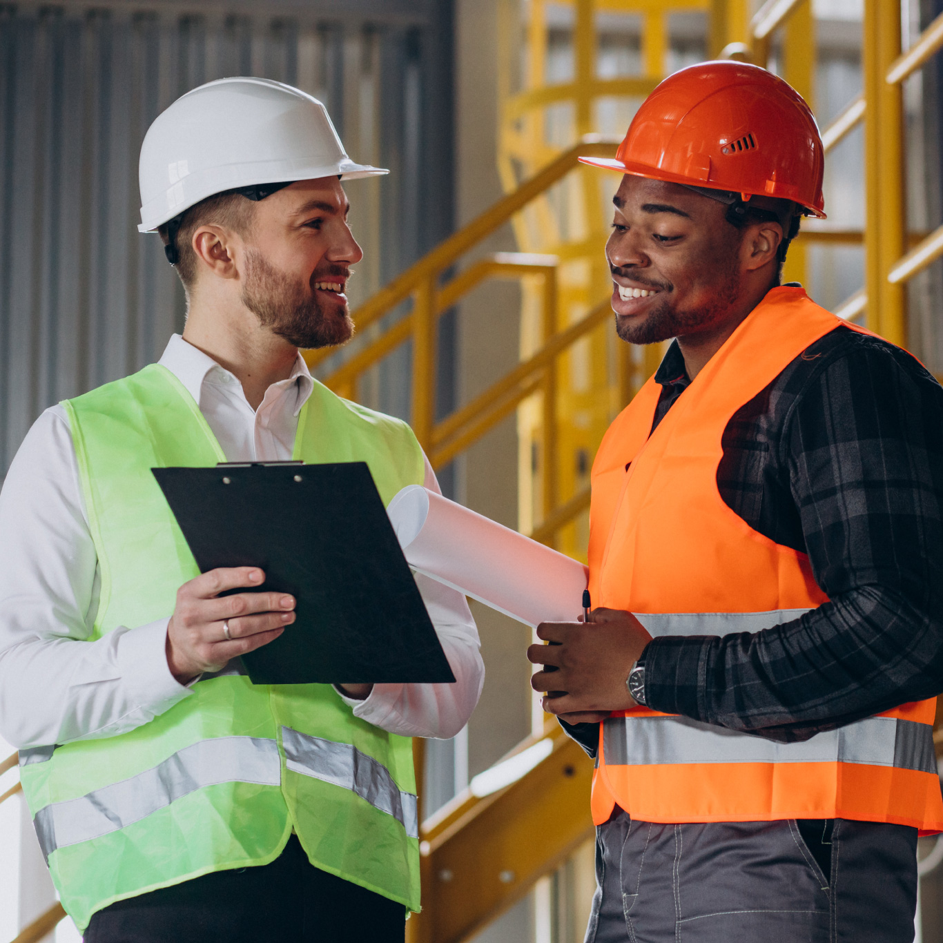Two male construction workers wearing hard hats and high-vis vests talking and looking at a clipboard