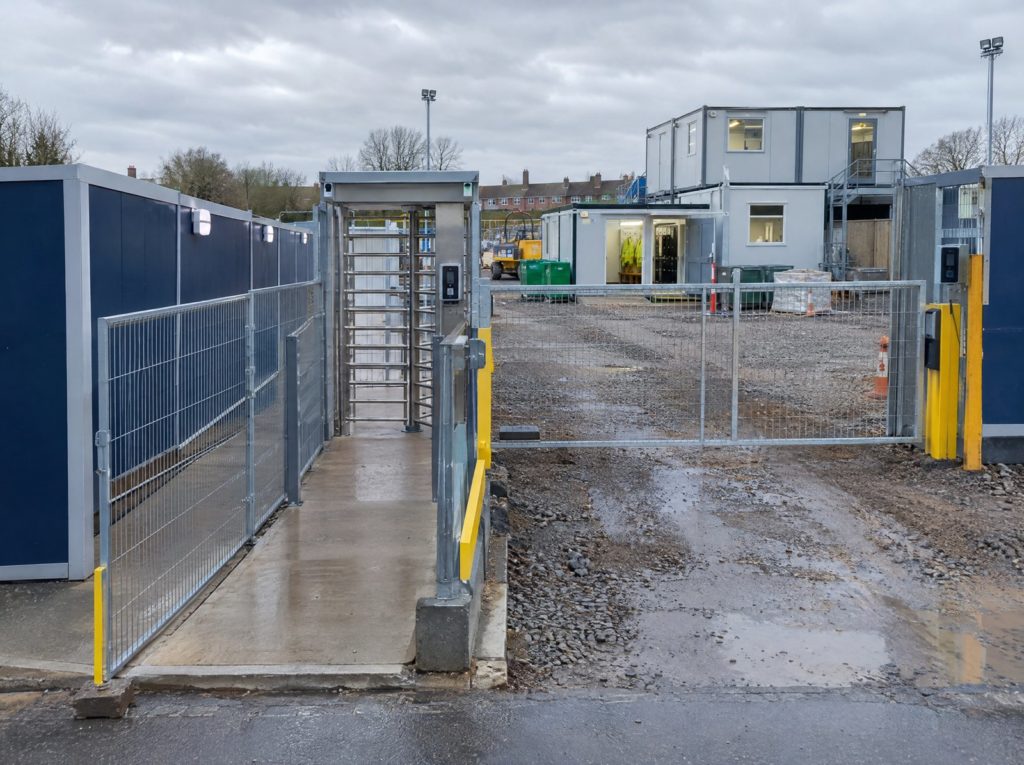 Construction site entrance with secure fencing, a turnstile, separate pedestrian walkway and welfare cabin
